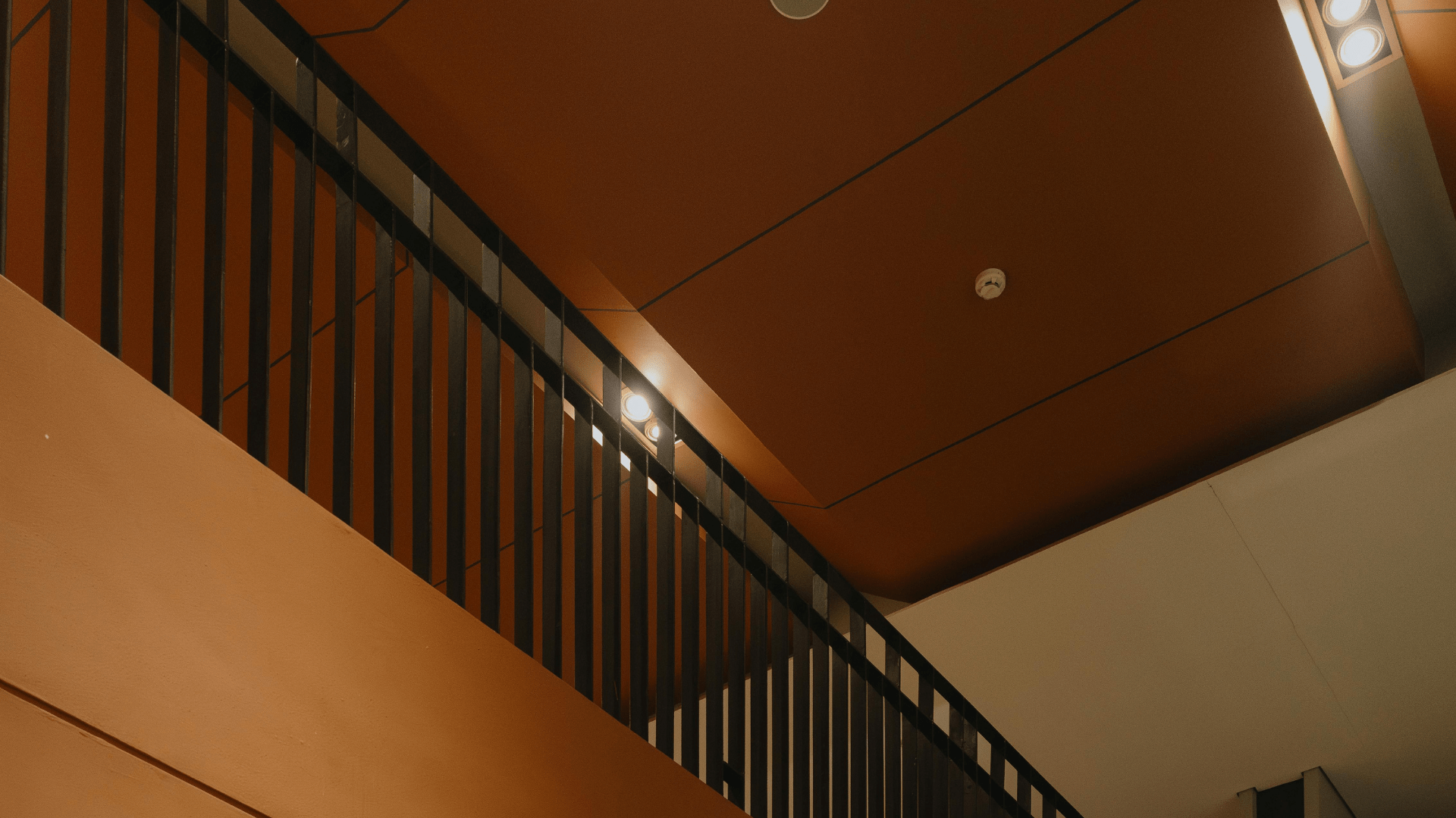 Barandilla interior de acero negro con barrotes verticales en escalera moderna Barandilla metálica negra con barrotes verticales en una escalera interior, con techo naranja y focos.